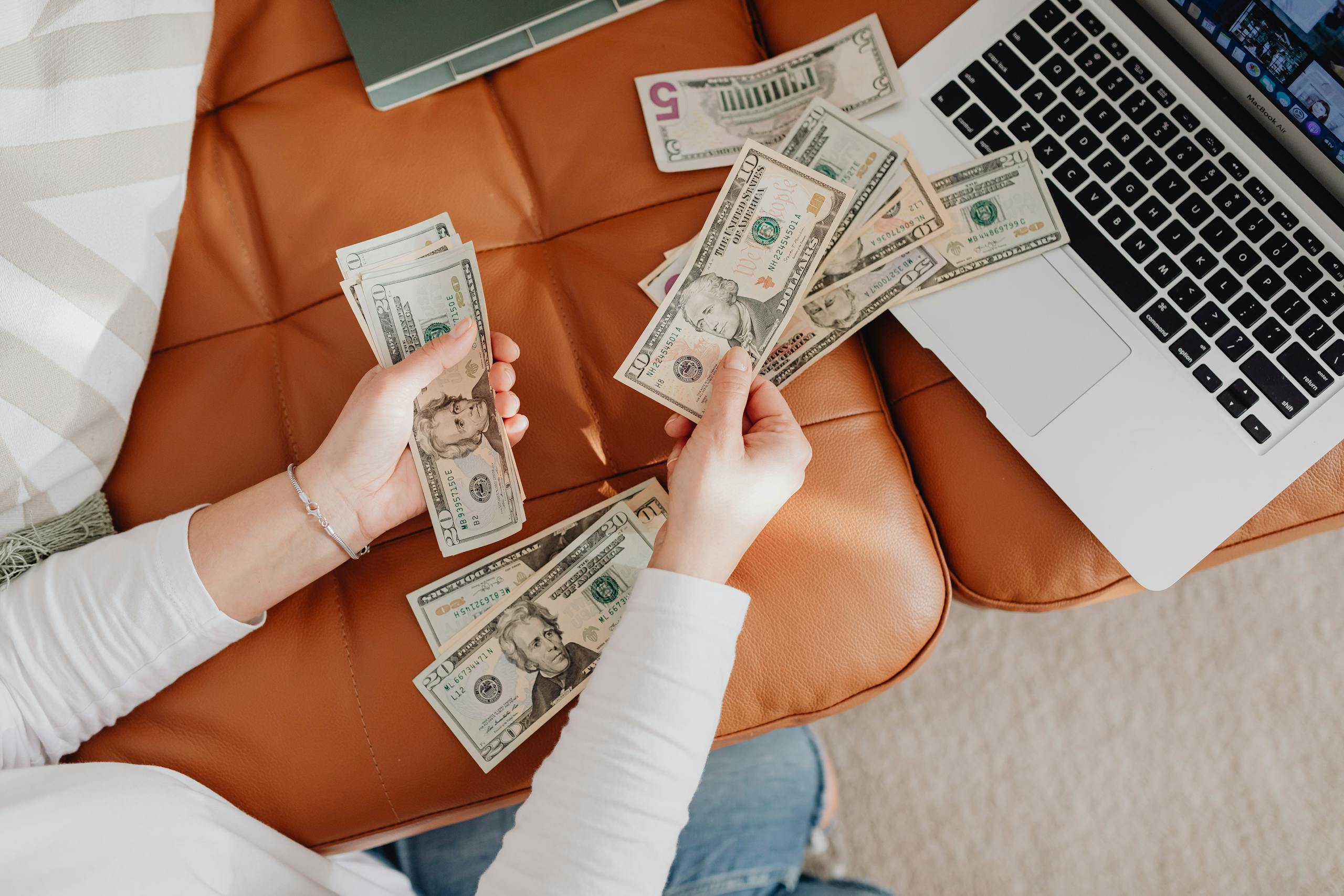 A person counts US dollar bills on a couch with a laptop, symbolizing financial planning.