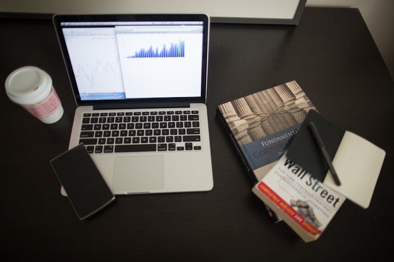 Top view of a desk with laptop, charts, books, and coffee, depicting a modern financial workspace setup.