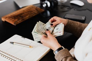 Unrecognizable elegant female in sweater counting dollar bills while sitting at wooden table with planner and pen